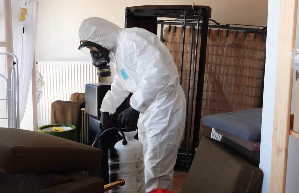 echnicien de DebugPro en combinaison de protection pulvérisant des produits chimiques dans une pièce pour éliminer les nuisibles, illustrant une intervention professionnelle et sécurisée.